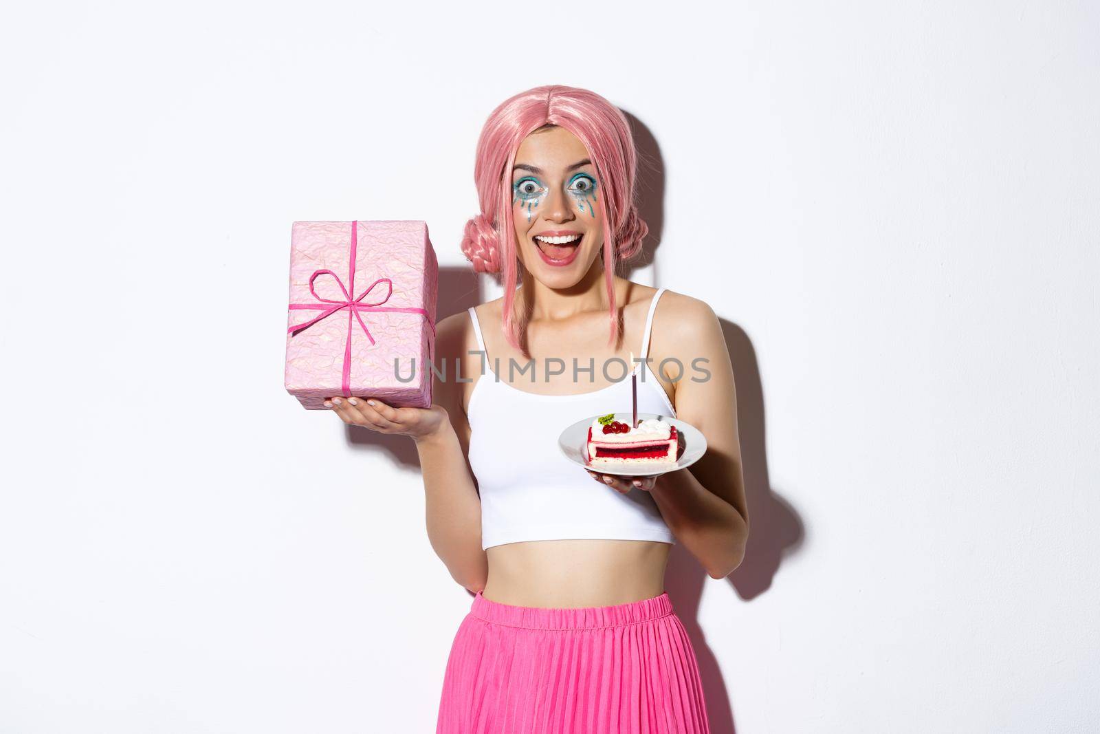 Portrait of surprised beautiful girl in pink wig, receive birthday gift, holding b-day cake and smiling happy, standing over white background.