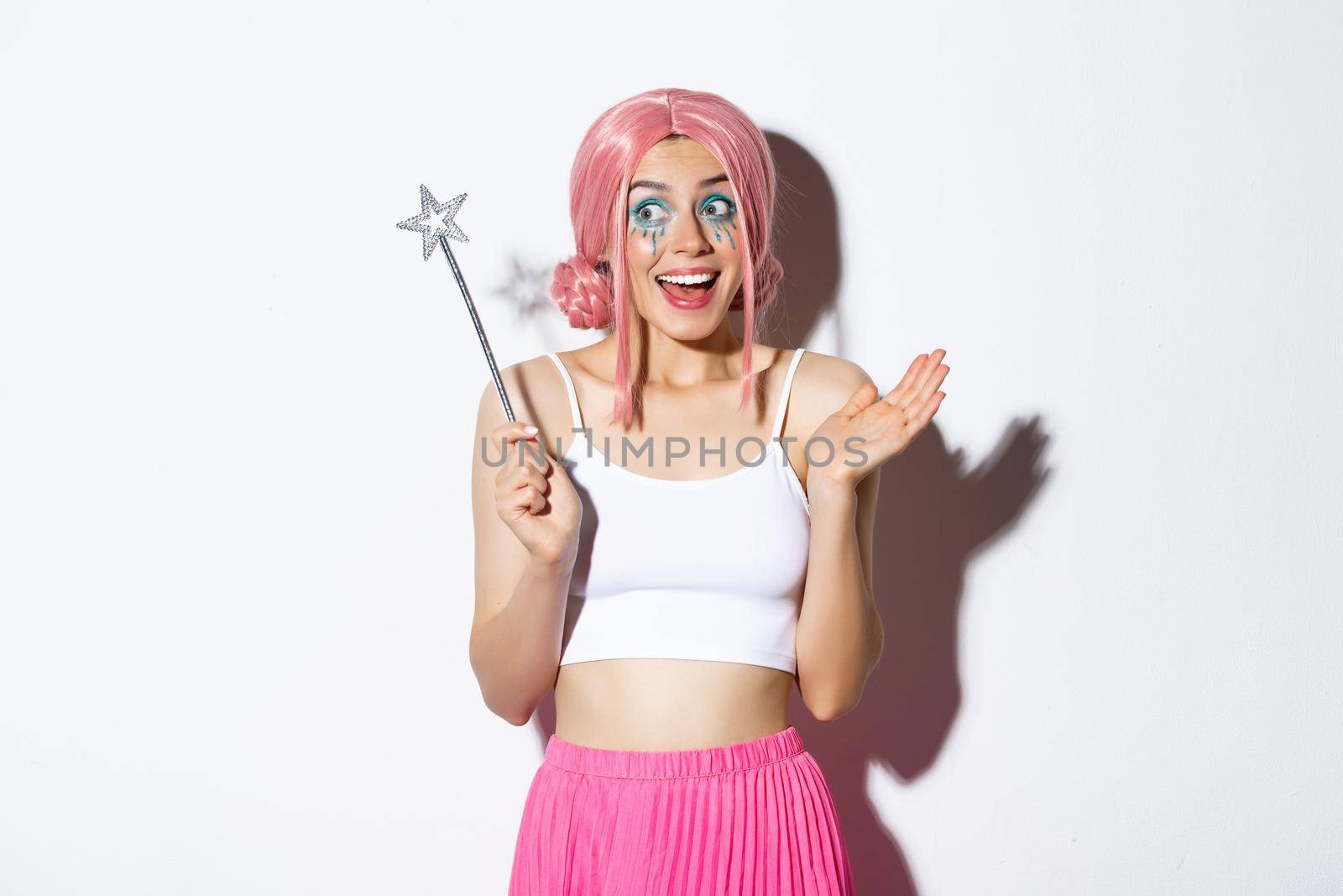 Portrait of pretty girl dressed up as cute fairy for halloween party, holding magic want and looking excited, celebrating holiday, standing over white background.