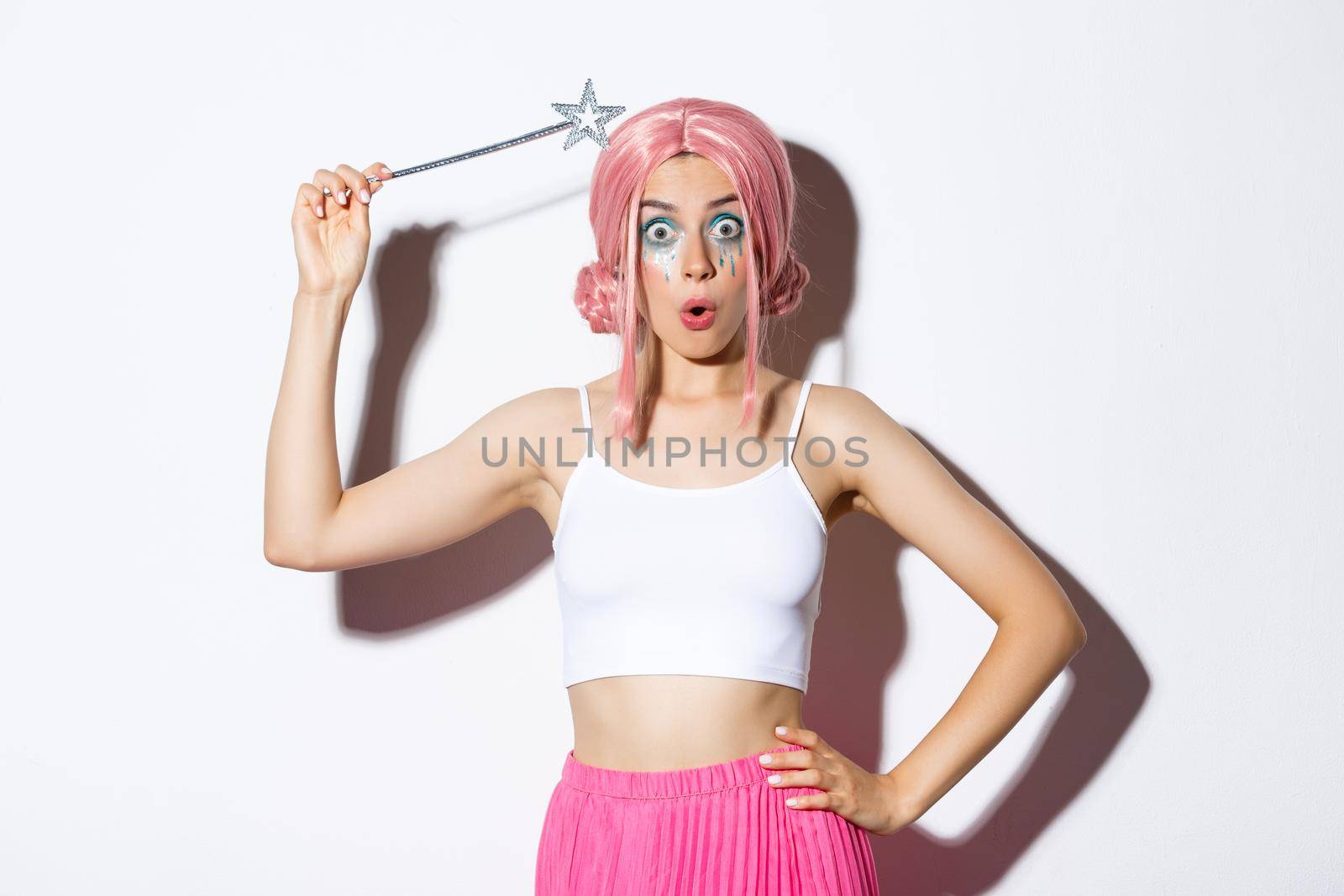 Image of silly attractive girl in pink wig, dressing as fairy for halloween party, hoding magic wand, standing over white background.