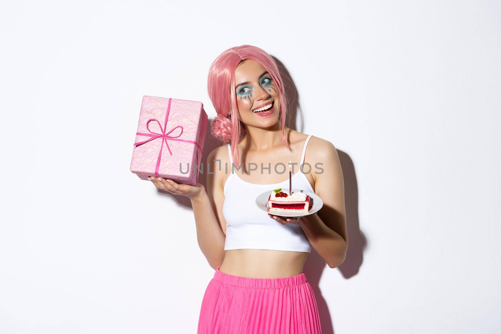 Image of excited cute girl in pink wig, shaking box with gift and wander what inside, holding piece of birthday cake, celebrating b-day.