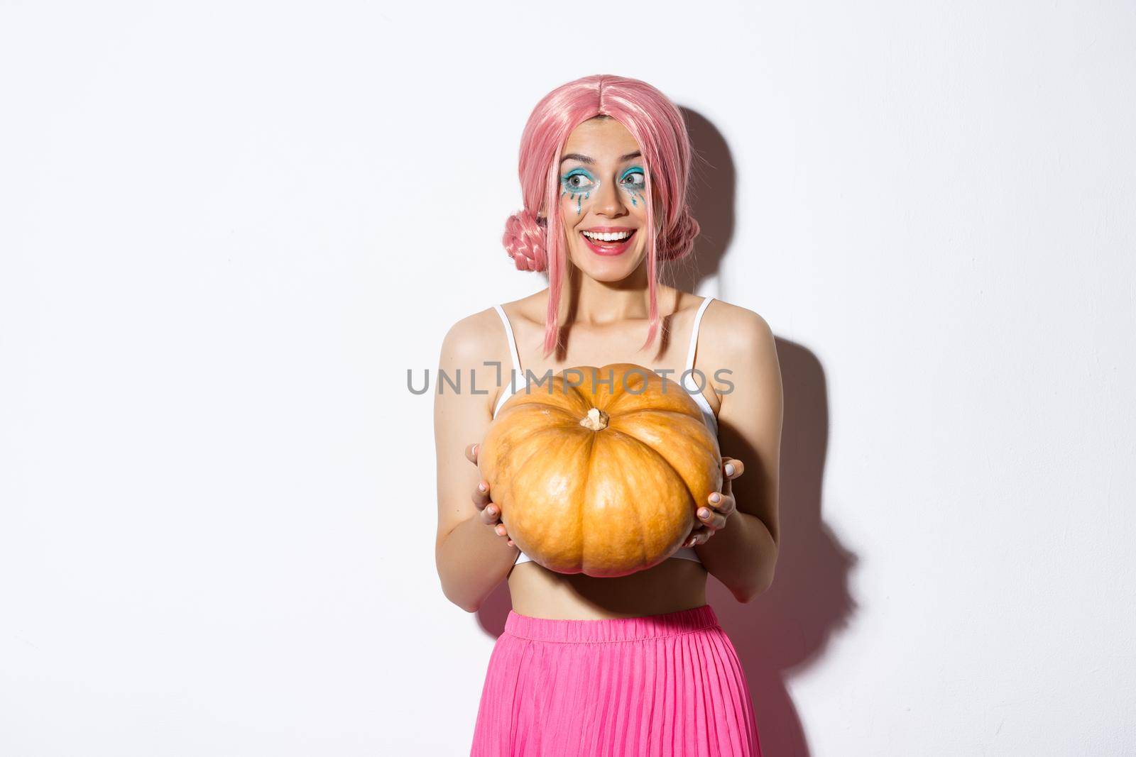 Portrait of attractive girl in pink wig looking amused and smiling, holding pumpkin for halloween party, standing over white background.