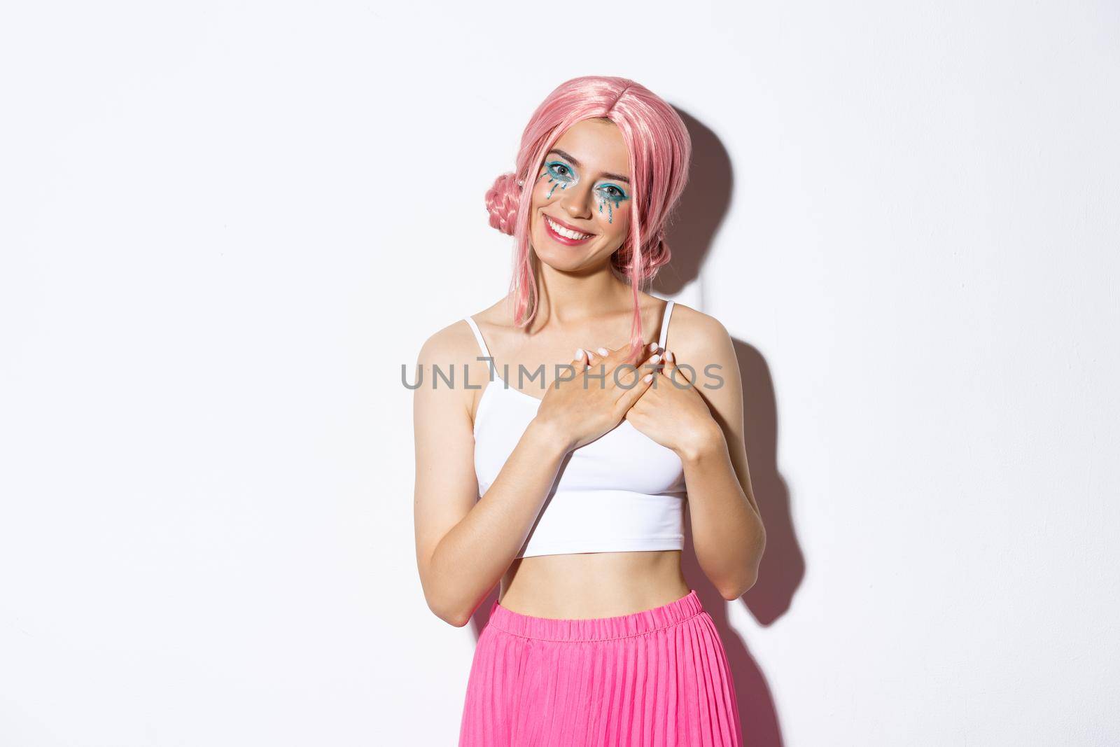 Image of cute party girl in pink wig looking thankful, holding hands on heart and smiling satisfied, standing against white background.