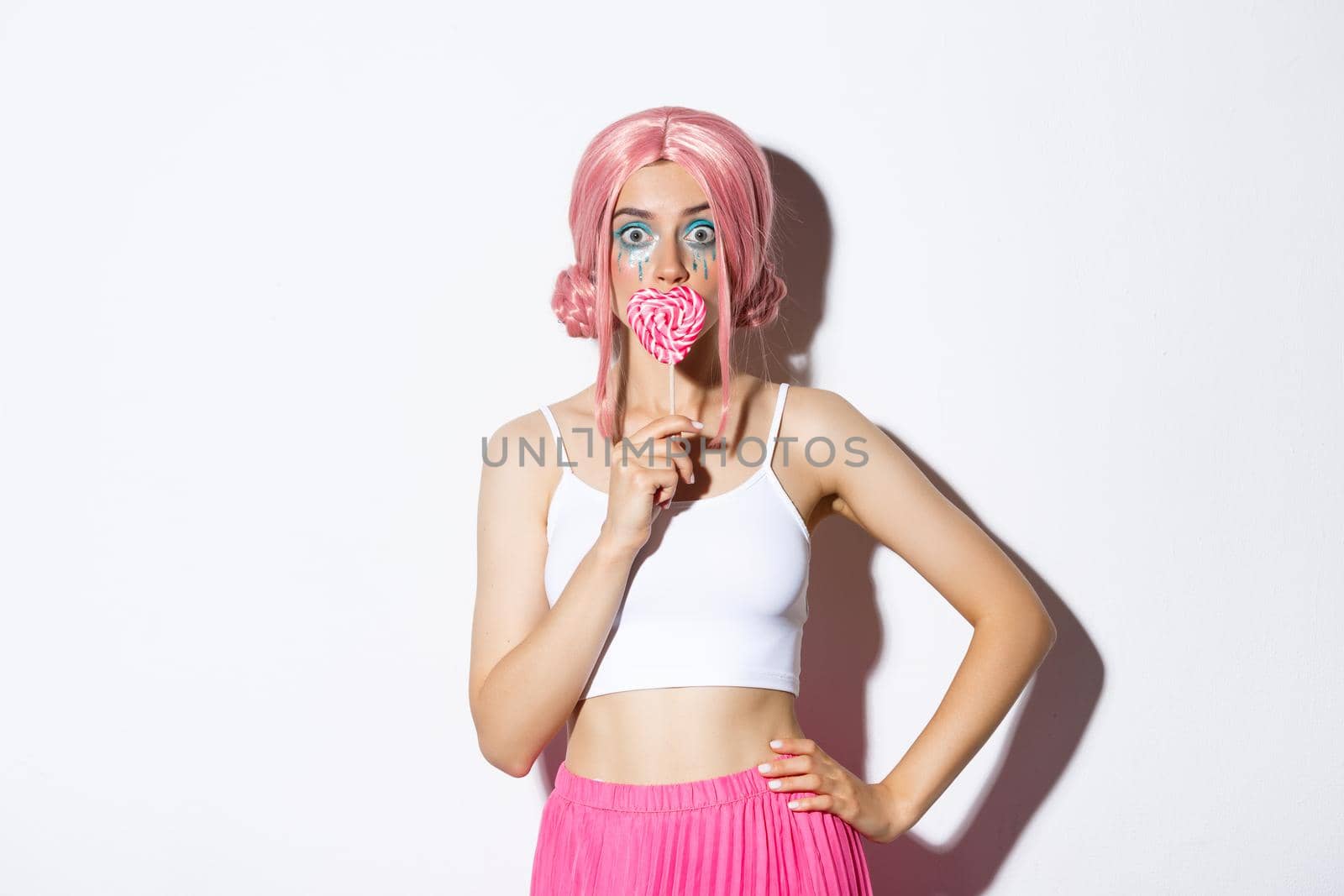 Beautiful smiling girl in pink wig, holding heart-shaped candy, trick or treating in fairy costume on halloween, standing over white background.