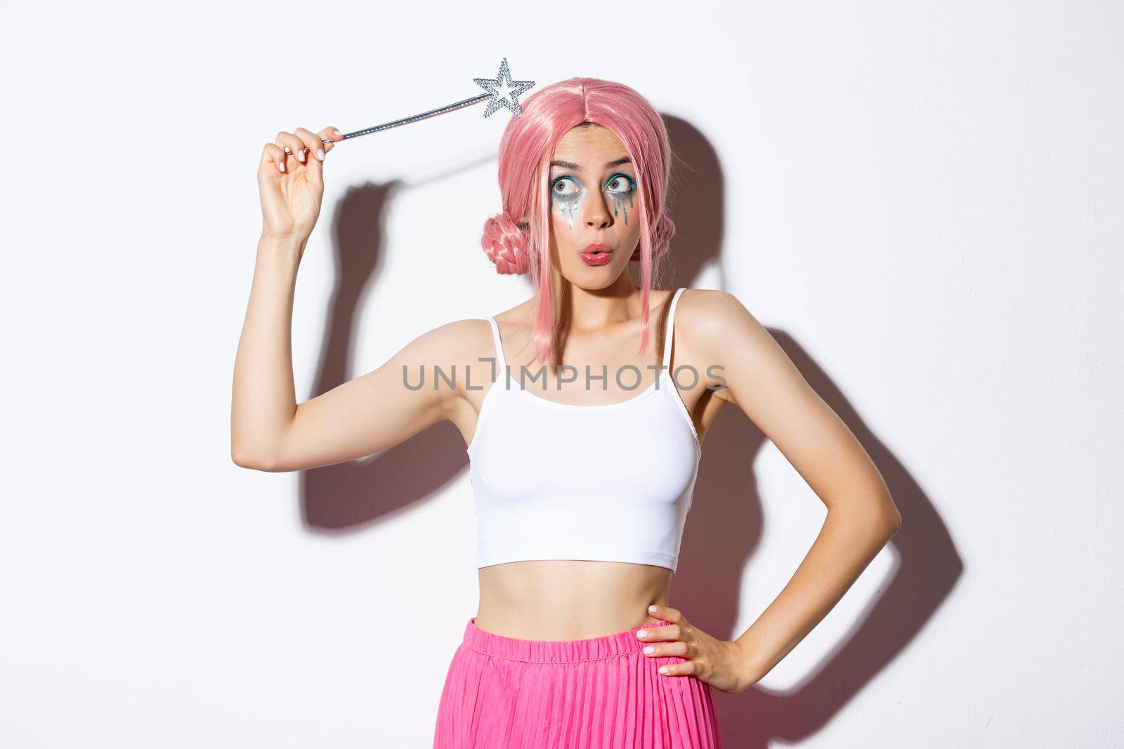 Image of silly attractive girl in pink wig, dressing as fairy for halloween party, hoding magic wand, standing over white background.