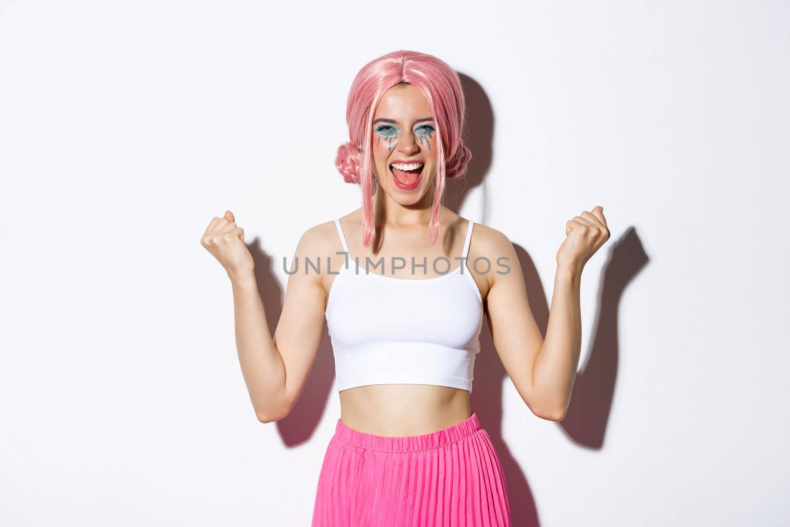 Image of excited successful girl in pink wig, celebrating something, making fist pump and smiling satisfied, enjoying halloween party, standing over white background.