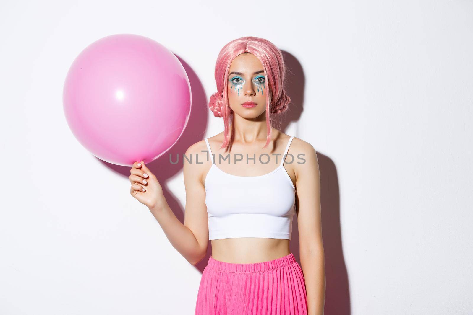 Image of young stylish girl holding pink balloon, wearing party wig and celebrating holiday, standing over white background.