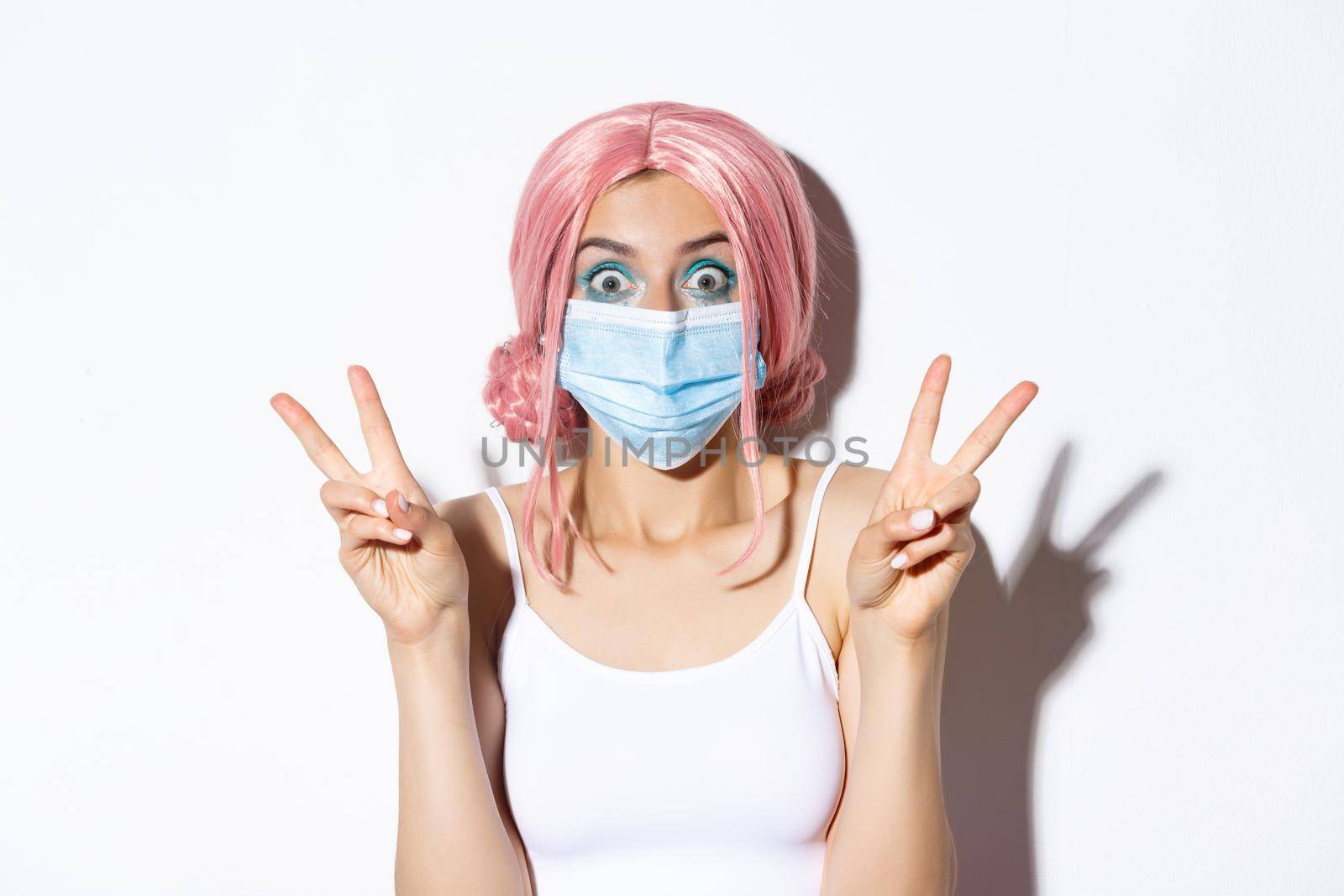 Coronavirus, social distancing and lifestyle concept. Close-up of pretty party girl in medical mask and pink wig, showing peace signs looking excited, white background.