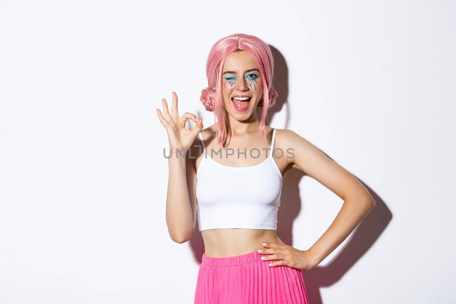 Beautiful cheeky girl in halloween costume and pink wig, showing okay sign in approval and winking, assure or recommend something, standing over white background.