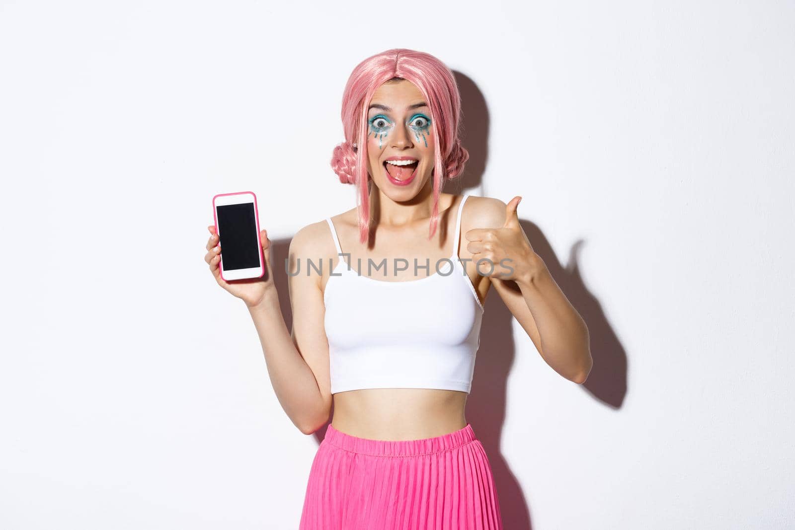 Excited stylish girl in pink wig, showing thumbs-up and looking fascnated while showing something awesome on mobile phone screen, standing over white background by Benzoix