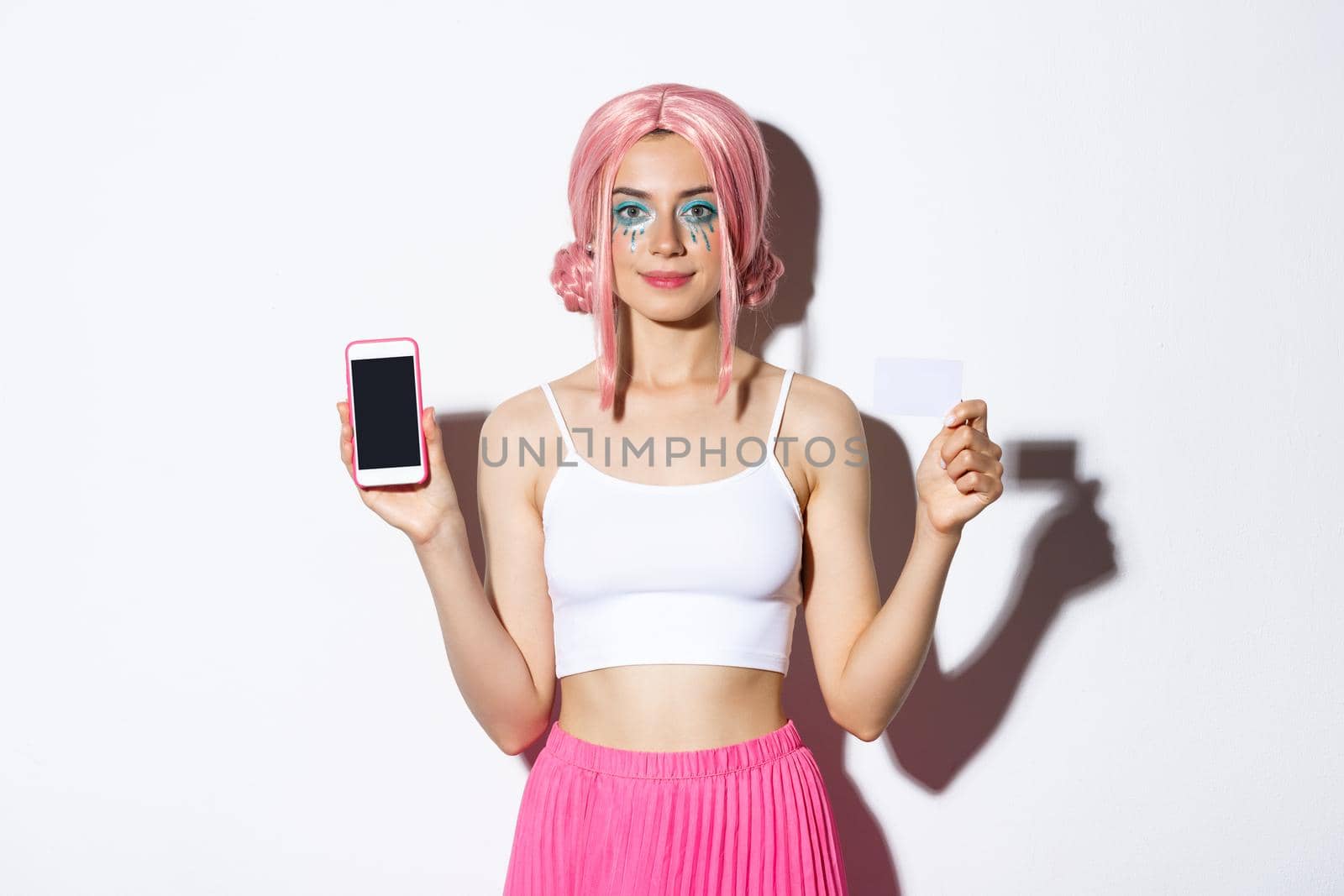 Image of stylish party girl in pink wig, showing credit card and mobile phone screen, celebrating holiday in glamour outfit, standing over white background by Benzoix