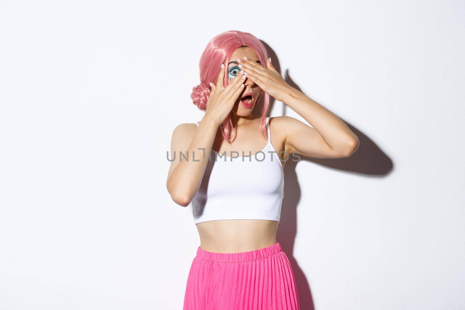 Portrait of silly pretty young woman in pink wig and halloween outfit, close eyes but peeking through fingers at something interesting.