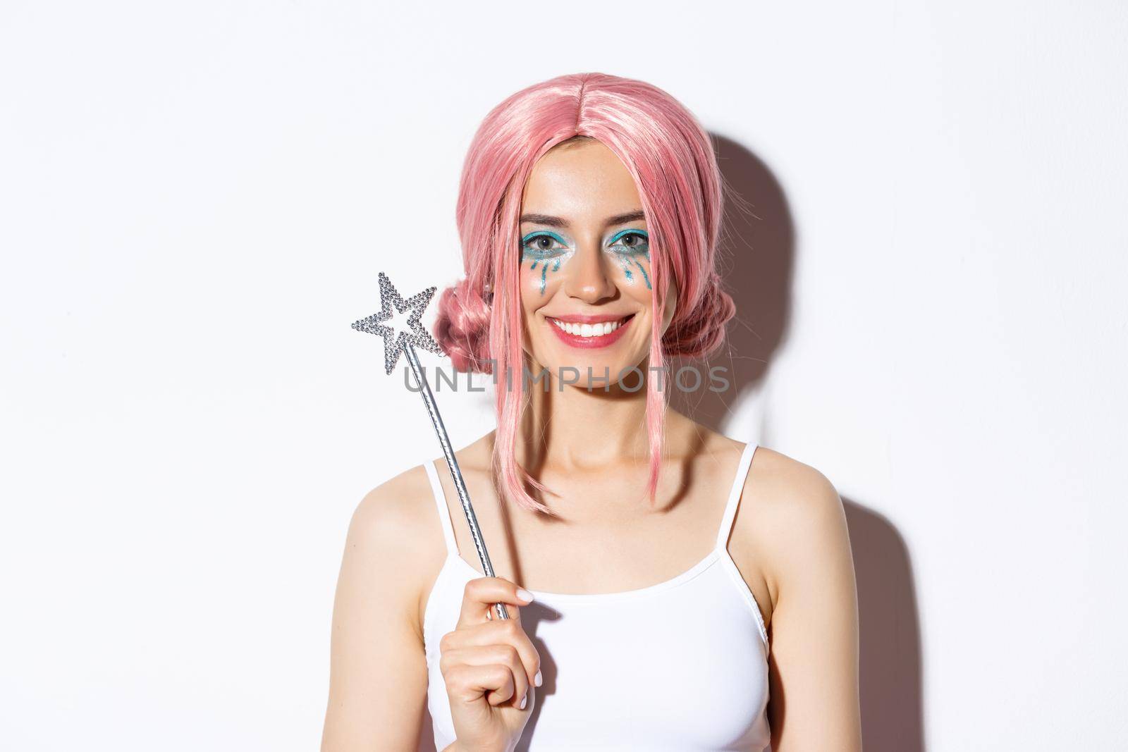 Close-up of beautiful girl dressed-up as a fairy for halloween party, holding magic wand and smiling, standing over white background by Benzoix