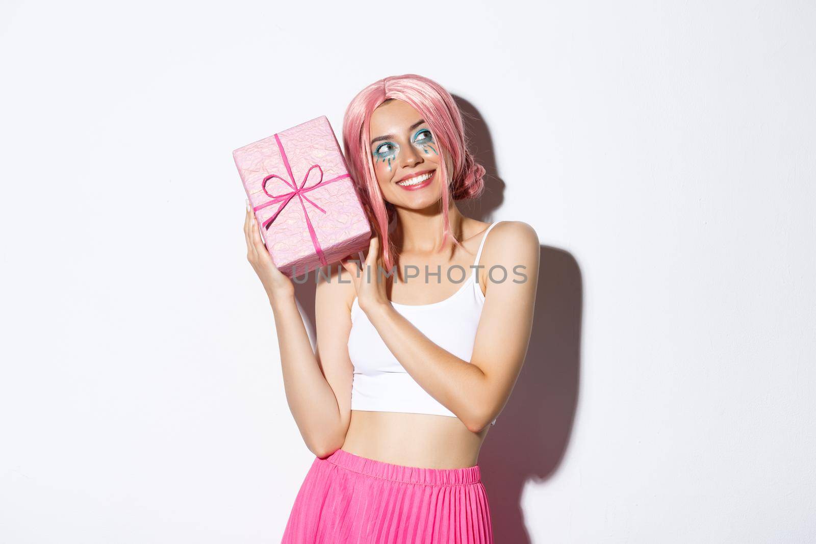 Image of beautiful girl in pink wig shaking box with birthday gift, wonder what inside wrapped box, standing over white background by Benzoix