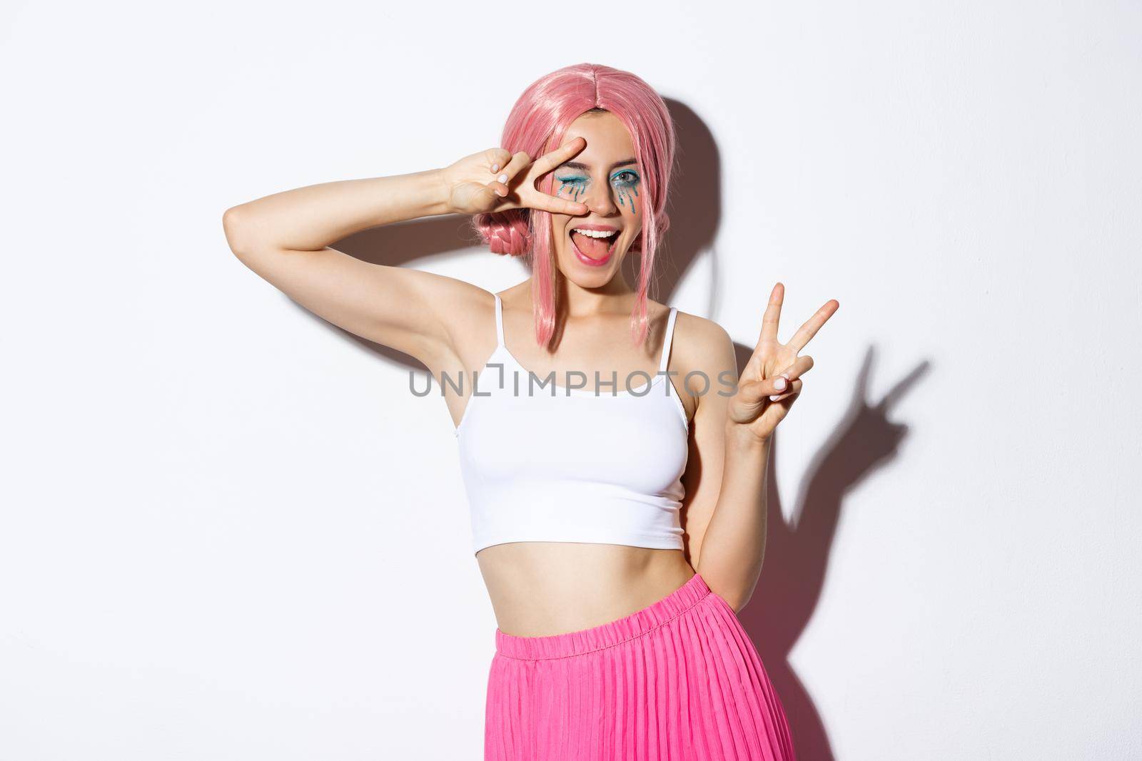 Happy beautiful girl in pink wig, dressed-up for halloween party, showing peace signs, smiling and winking coquettish at camera.