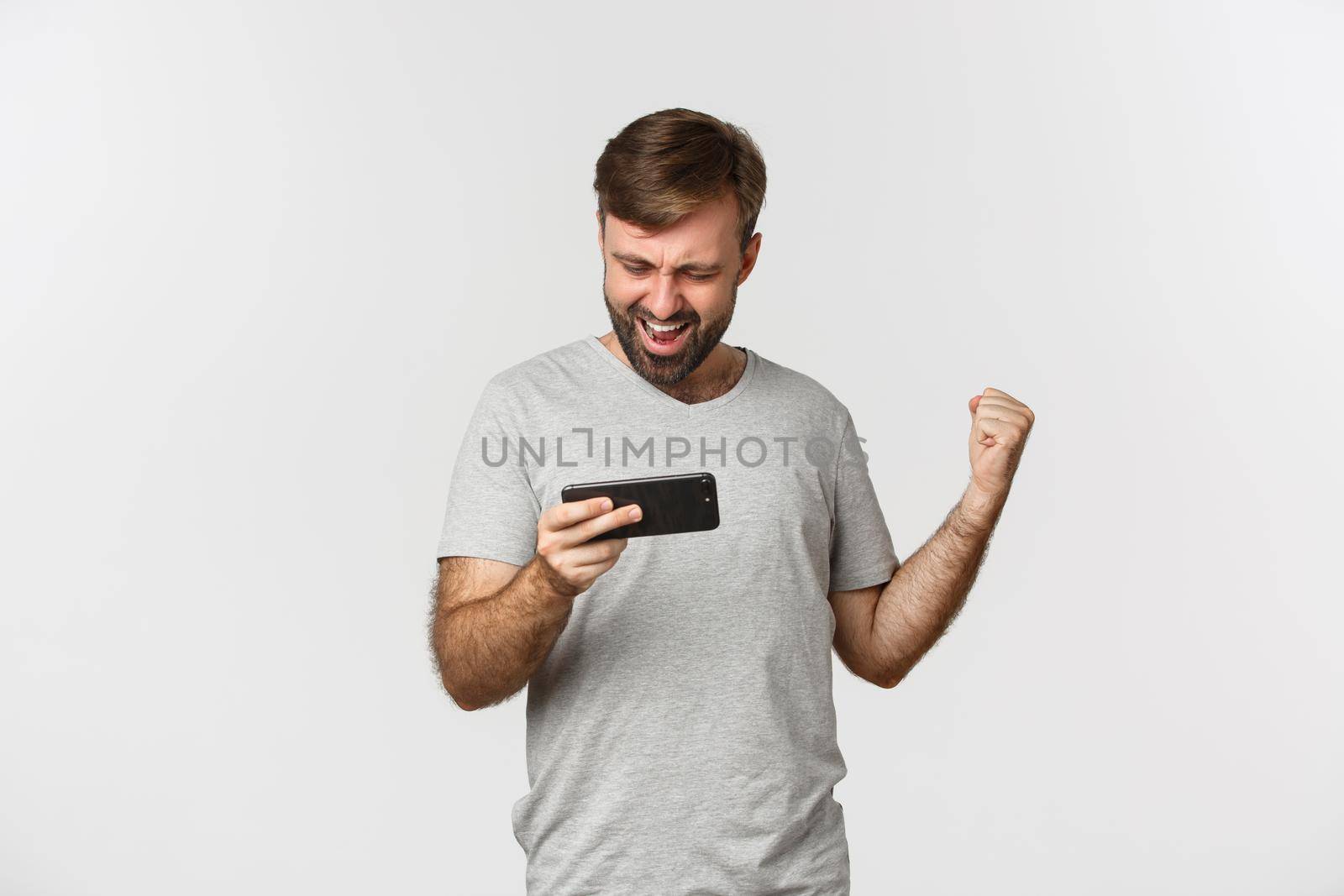 Image of adult bearded man in gray t-shirt, playing mobile games and winning, standing over white background.