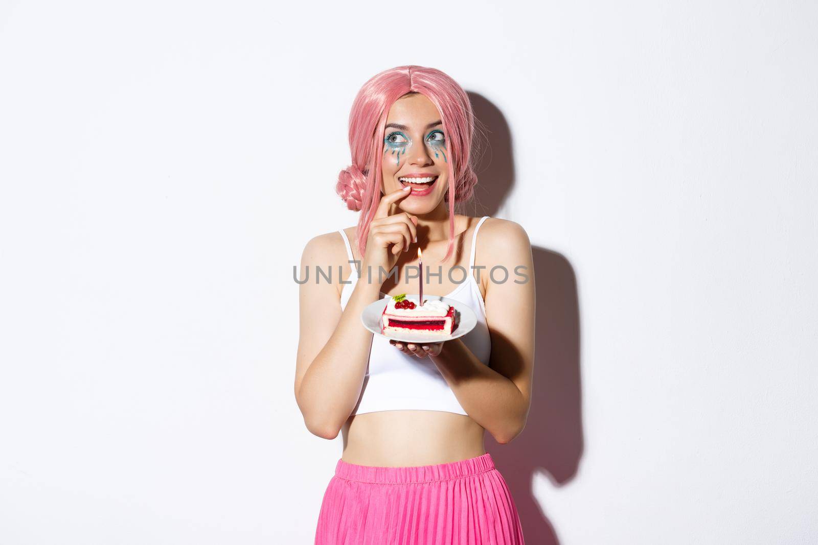 Portrait of lovely birthday girl in pink party wig, holding cake, thinking before making wish, standing over white background.