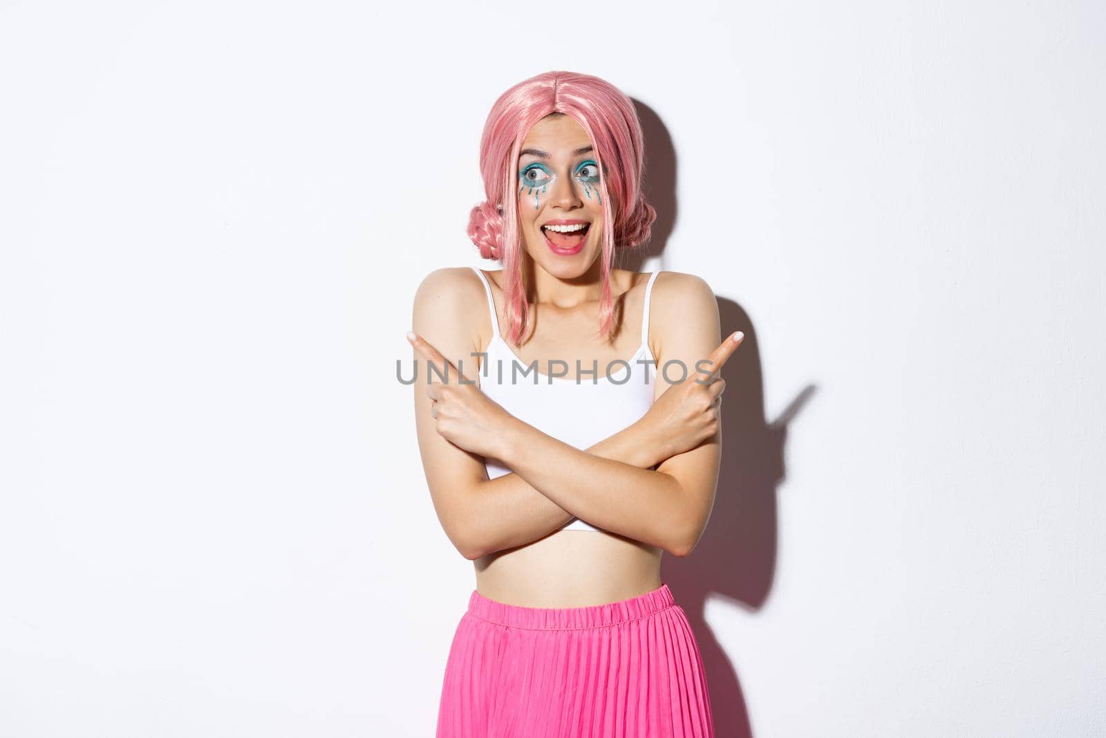 Portrait of excited smiling girl with pink wig and party makeup, pointing fingers sideways but looking left with happy expression, standing over white background by Benzoix