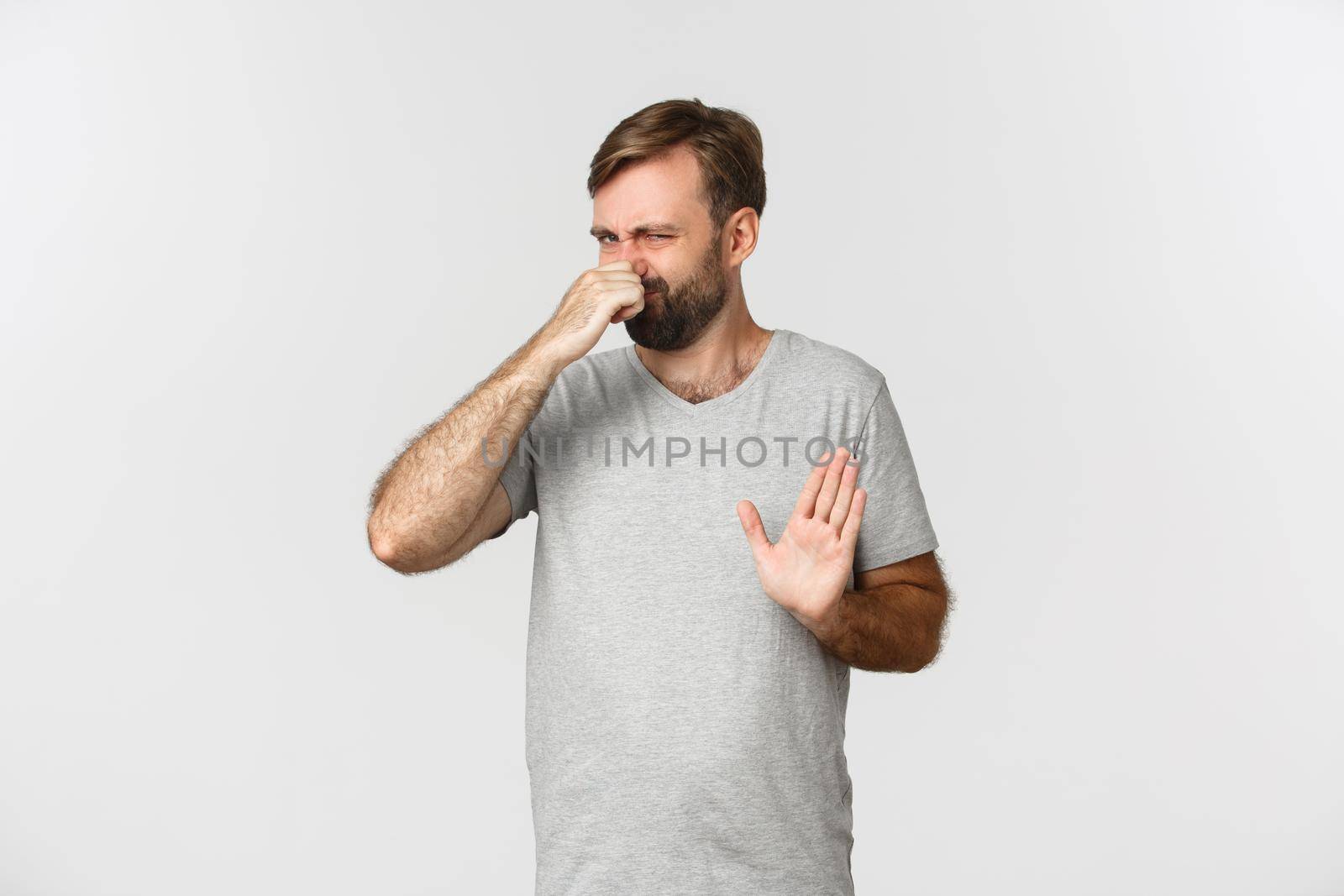 Image of an ordinary guy in gray t-shirt shut his nose, rejecting something disgusting with awful smell, standing over white background by Benzoix