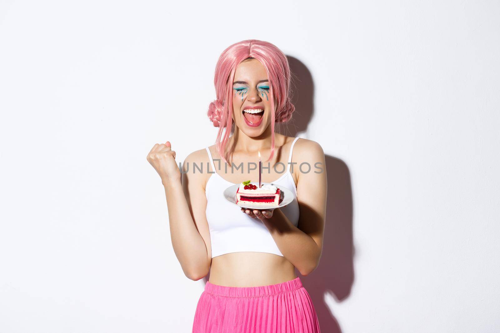 Portrait of cheerful smiling girl celebrating her birthday, wearing pink wig, holding b-day cake and shouting of joy, standing over white background.