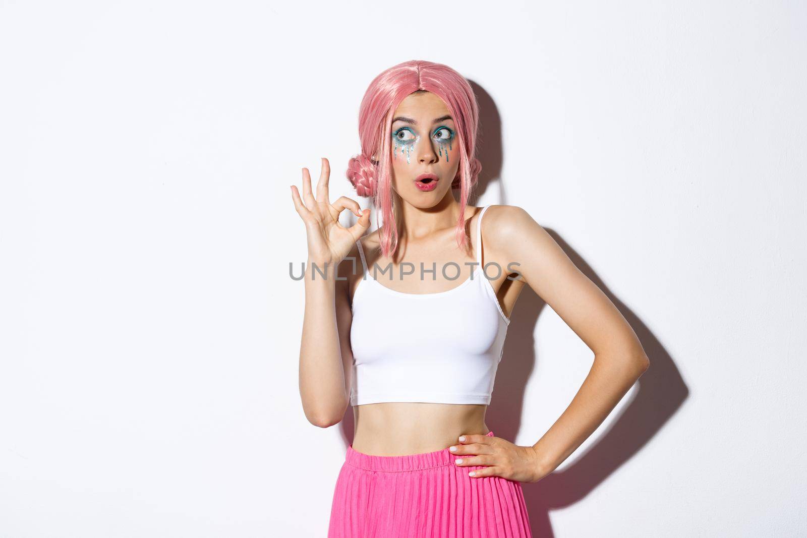 Portrait of coquettish party girl in pink wig and halloween outfit, showing okay sign and looking at upper left corner advertisement, standing over white background.
