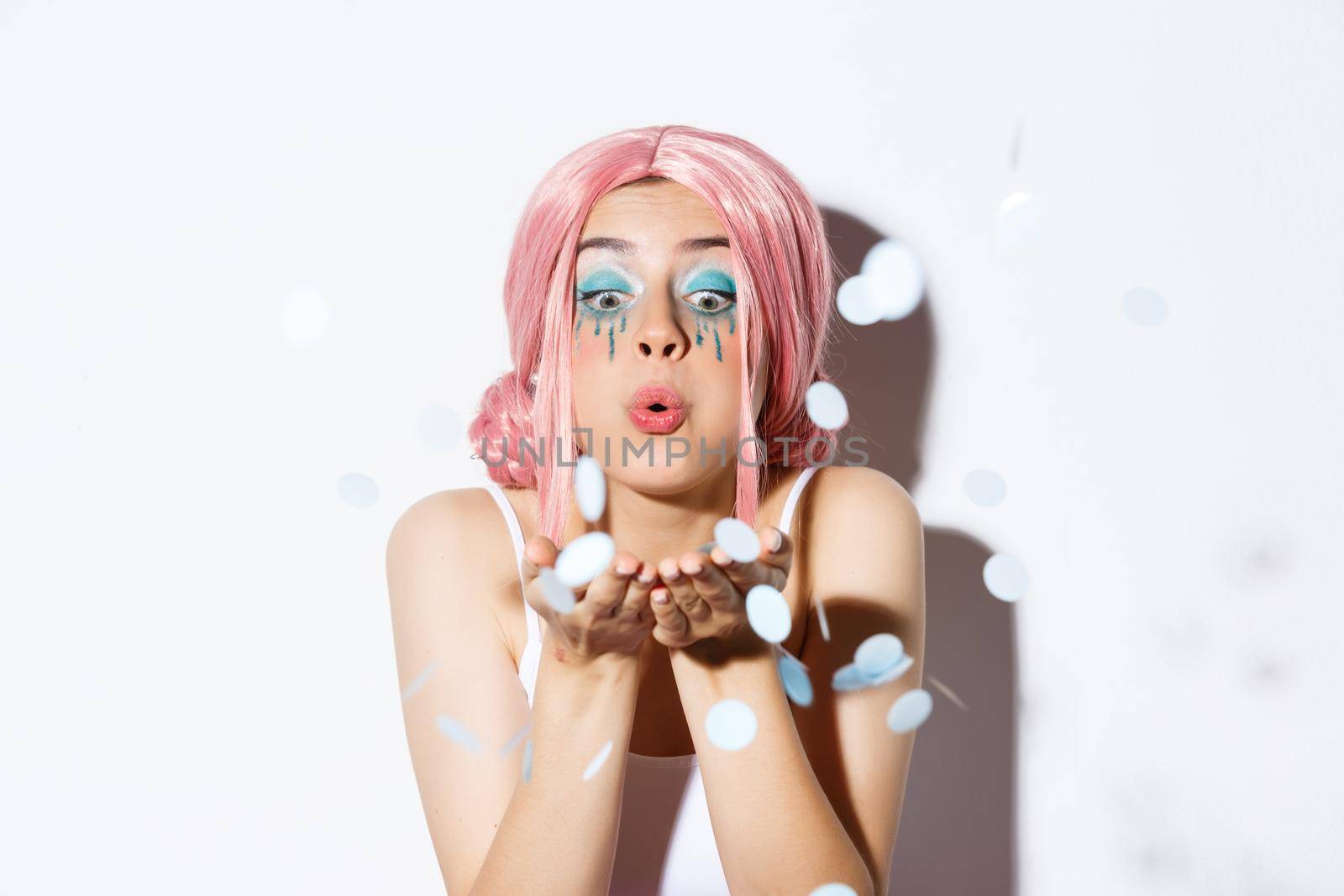 Close-up of lovely pretty party girl with pink wig and bright makeup, blowing confetti from palm, celebrating holiday, standing over white background by Benzoix