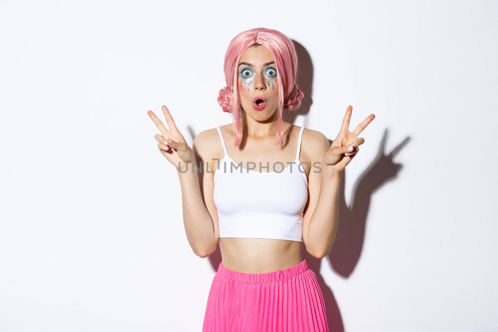 Portrait of amazed gasping girl looking at something fantastic, showing peace signs, wearing halloween costume for party, holiday celebration, standing over white background.