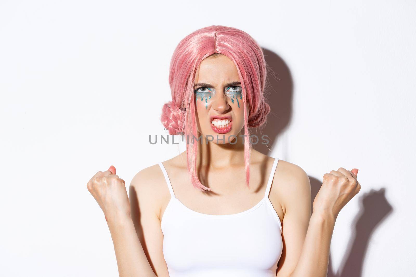 Close-up of angry distressed girl in pink wig, clenching fists and looking up with hatred, standing over white background.