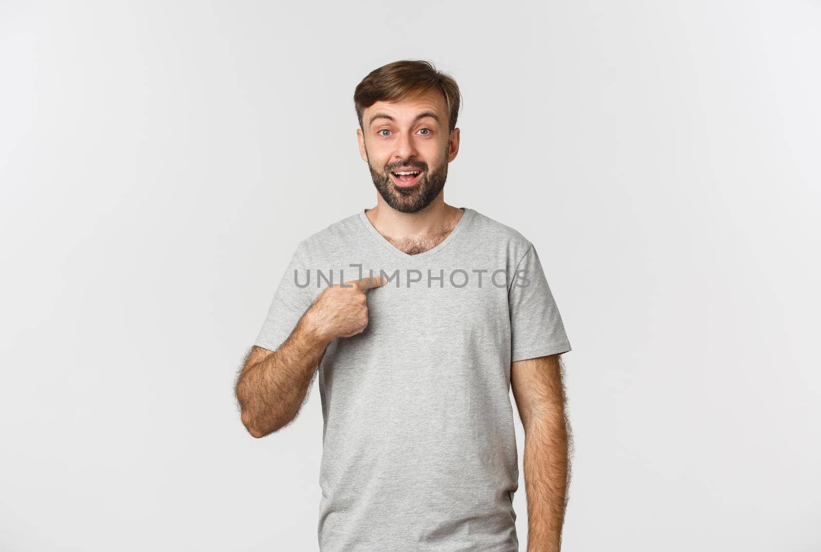 Image of happy and surprised man in gray t-shirt, pointing at himself, standing over white background.