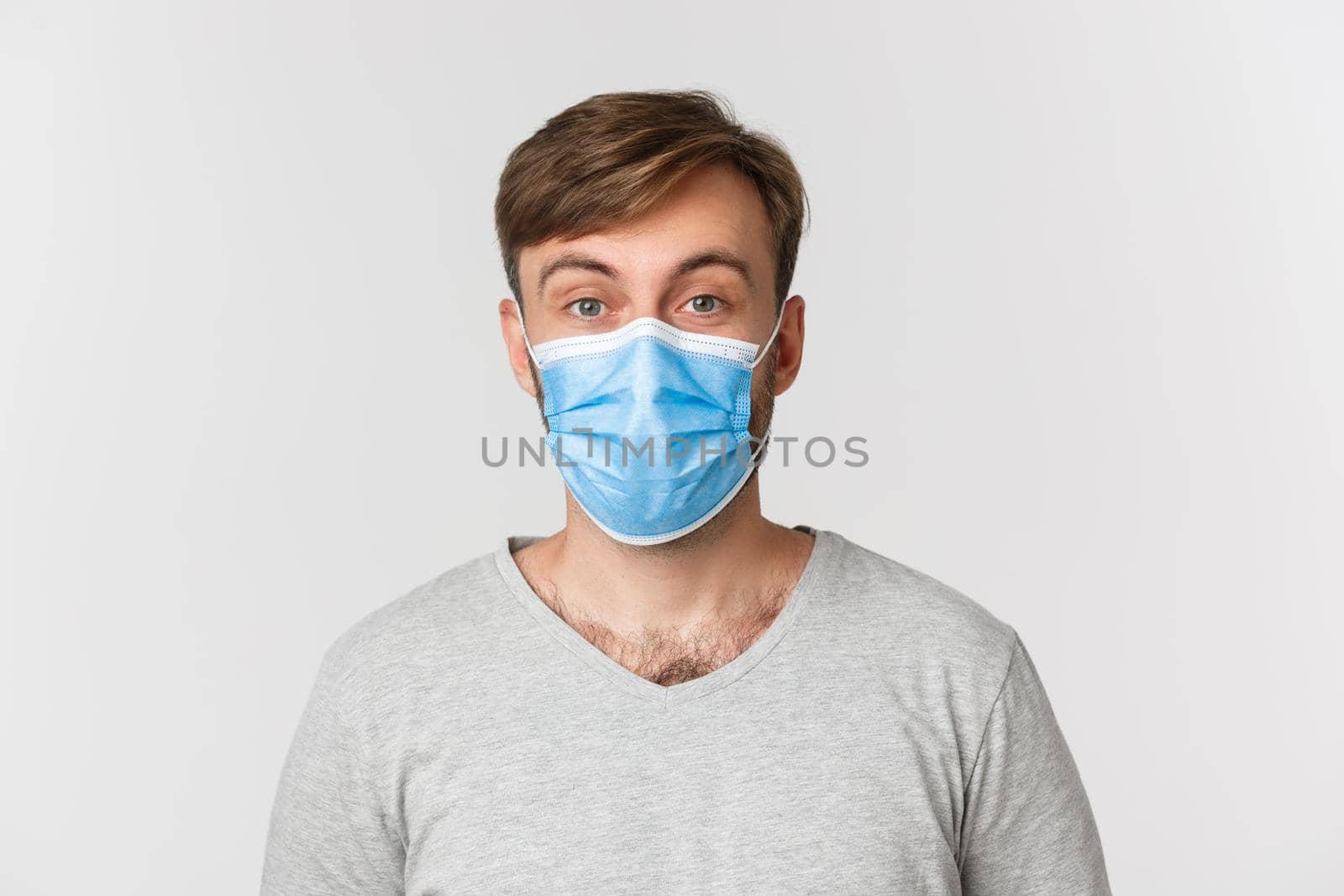Concept of pandemic, covid-19 and social-distancing. Close-up of surprised man in medical mask, raising eyebrows amazed, standing over white background.