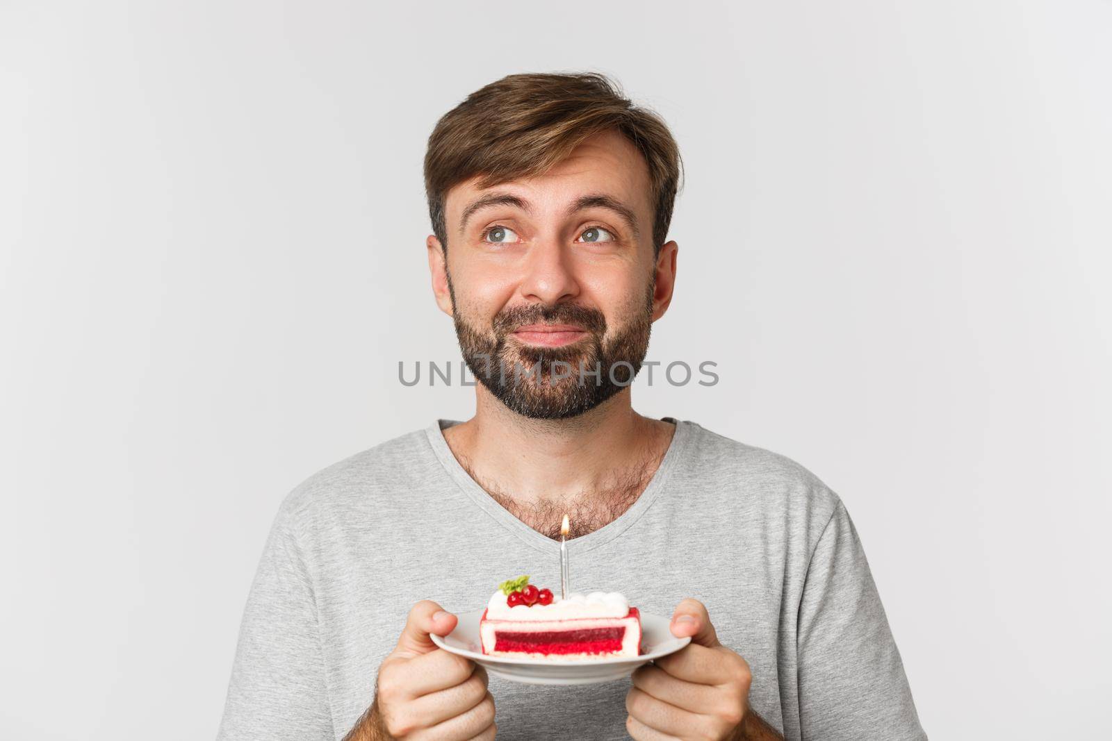 Close-up of dreamy bearded man, smiling and celebrating birthday, holding cake with lit candle, making b-day wish, standing over white background.