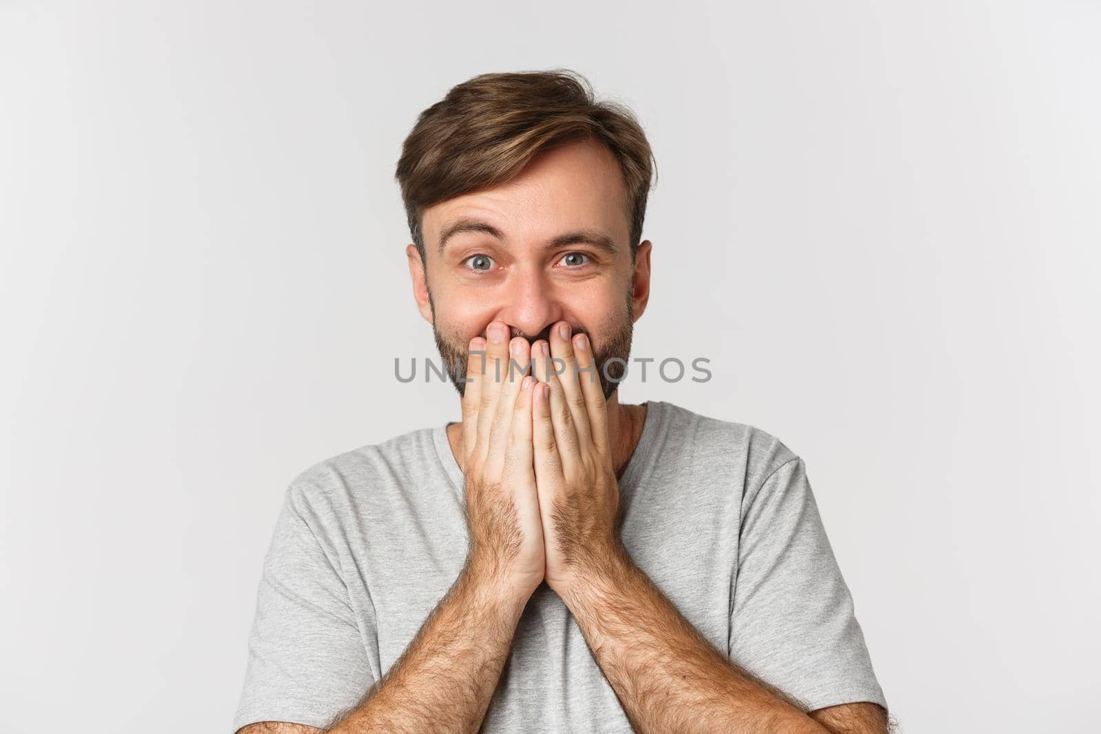 Close-up of excited and surprised man, cover mouth and giggle, standing over white background.