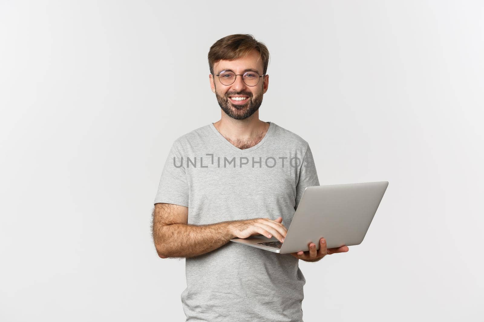 Young handsome freelancer in glasses and gray t-shirt, smiling pleased, working with laptop, standing over white background.