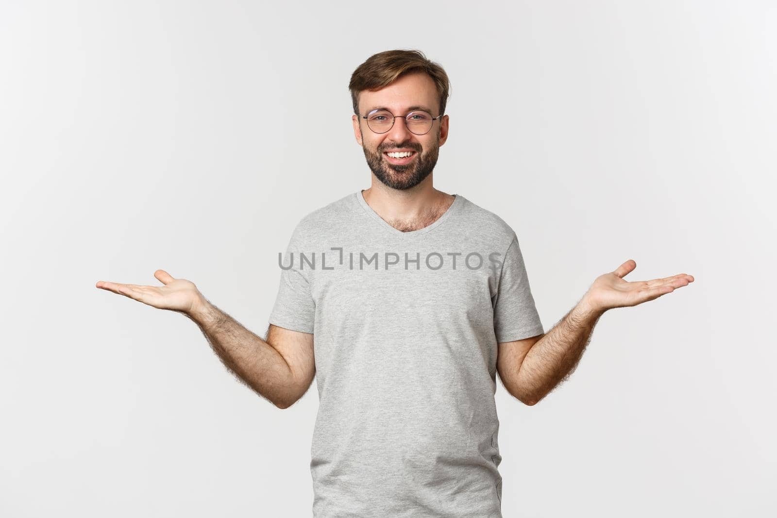 Handsome man with beard, wearing gray t-shirt and glasses, spread hands sideways and holding two things, demonstrate products, standing over white background.
