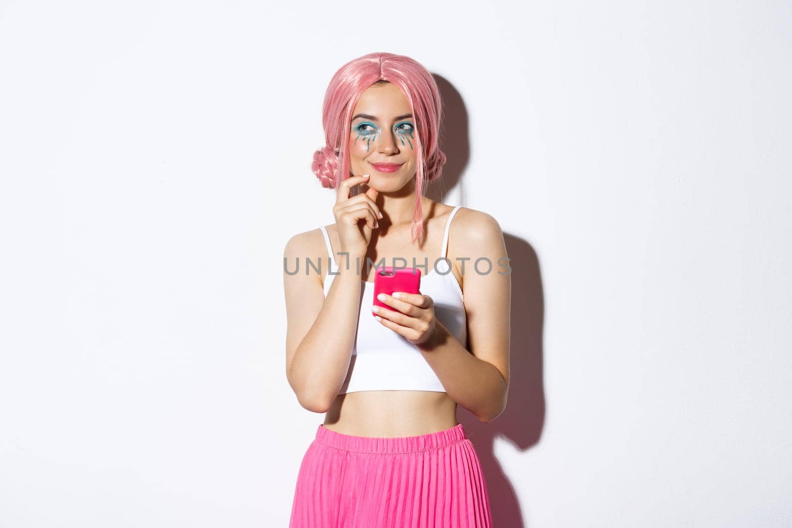 Portrait of thoughtful girl have interesting idea, looking away and thinking while holding smartphone, wearing pink wig for party, celebrating holiday.