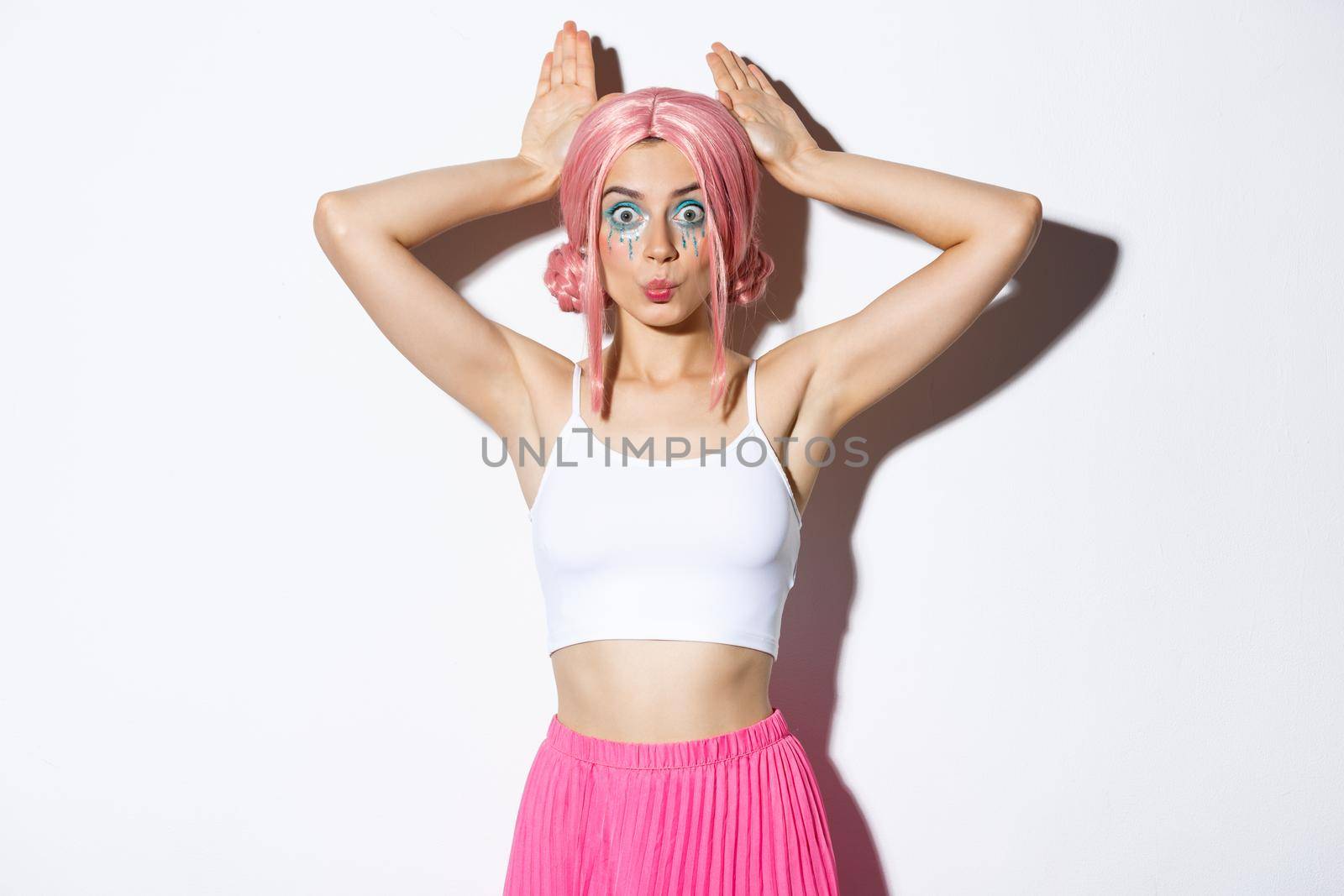 Portrait of lovely party girl in pink anime wig, showing bunny ears and pouting silly, celebrating halloween, standing over white background.