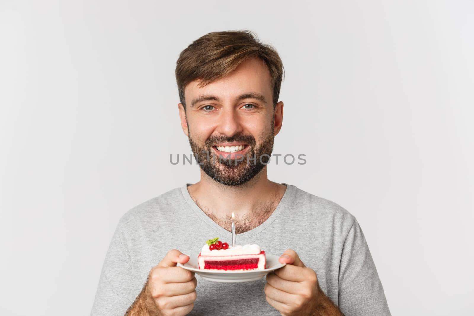 Close-up of happy bearded man, smiling and celebrating birthday, holding cake with lit candle, making b-day wish, standing over white background by Benzoix