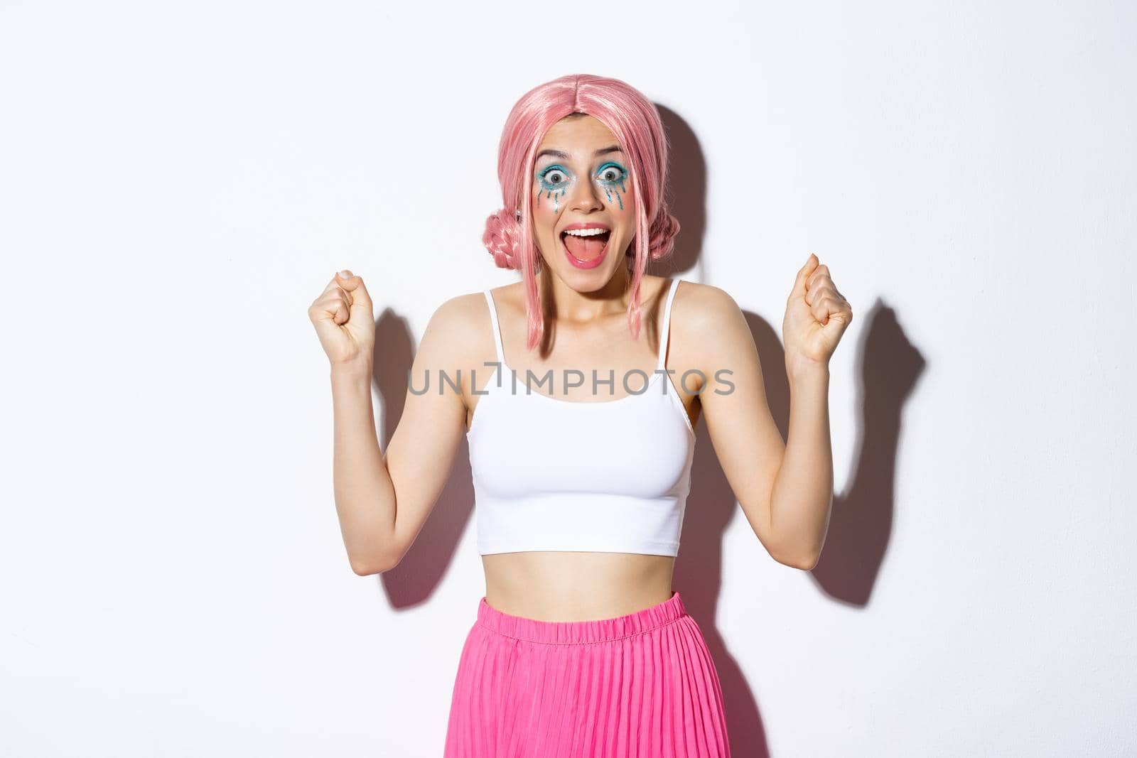 Image of cheerful girl in pink wig and halloween costume, raising hands up like a winner, celebrating victory, looking amazed and delighted, standing over white background.
