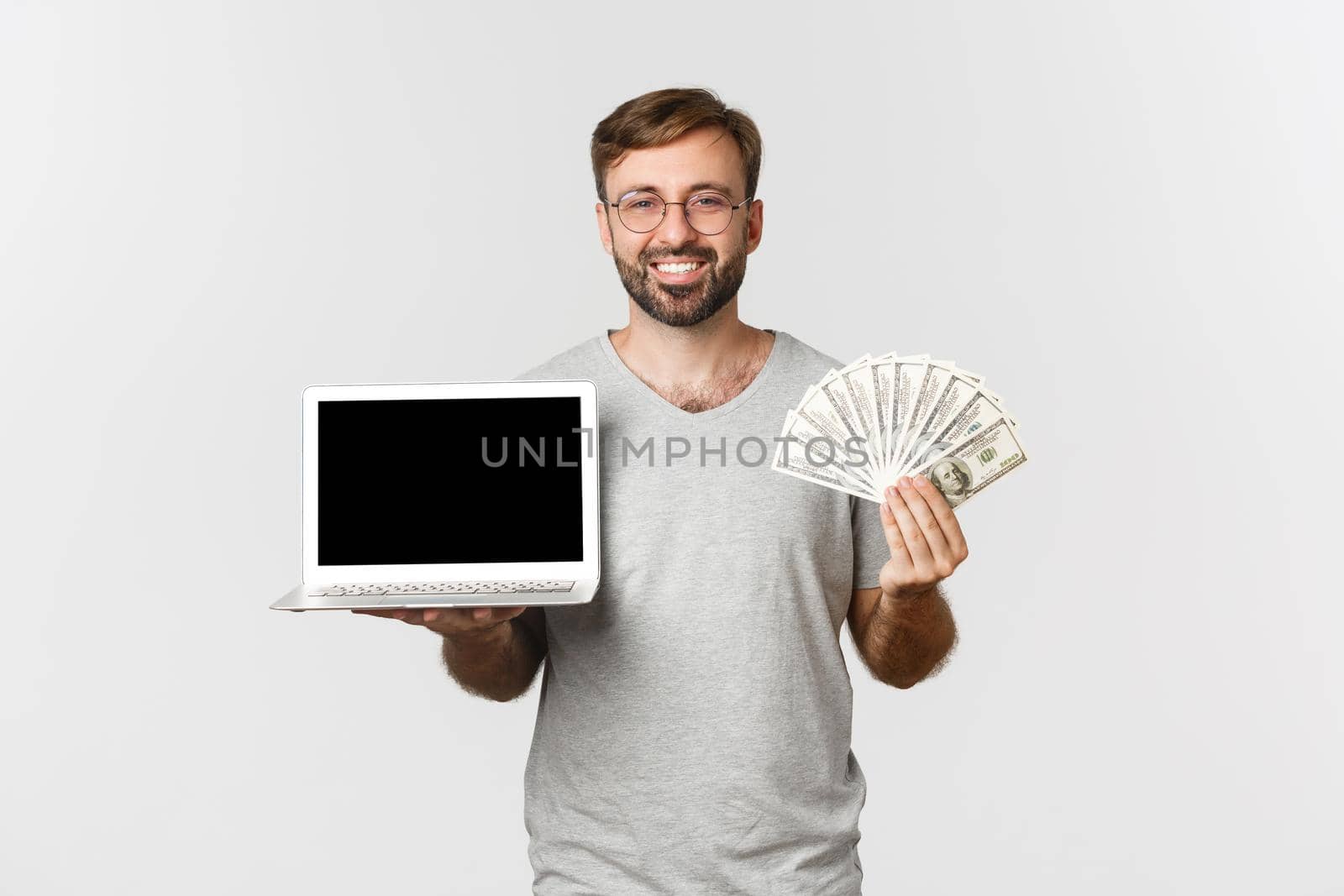 Image of handsome man with beard, working with laptop, networking, holding money and smiling, standing over white background.