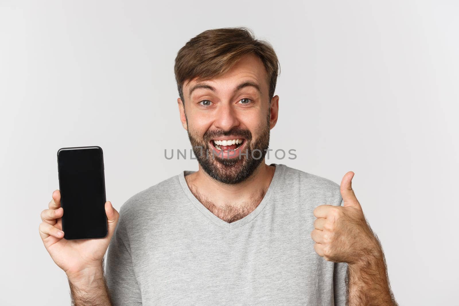 Close-up of smiling handsome man in gray t-shirt, showing mobile phone screen and thumbs-up, recommending app, standing over white background.