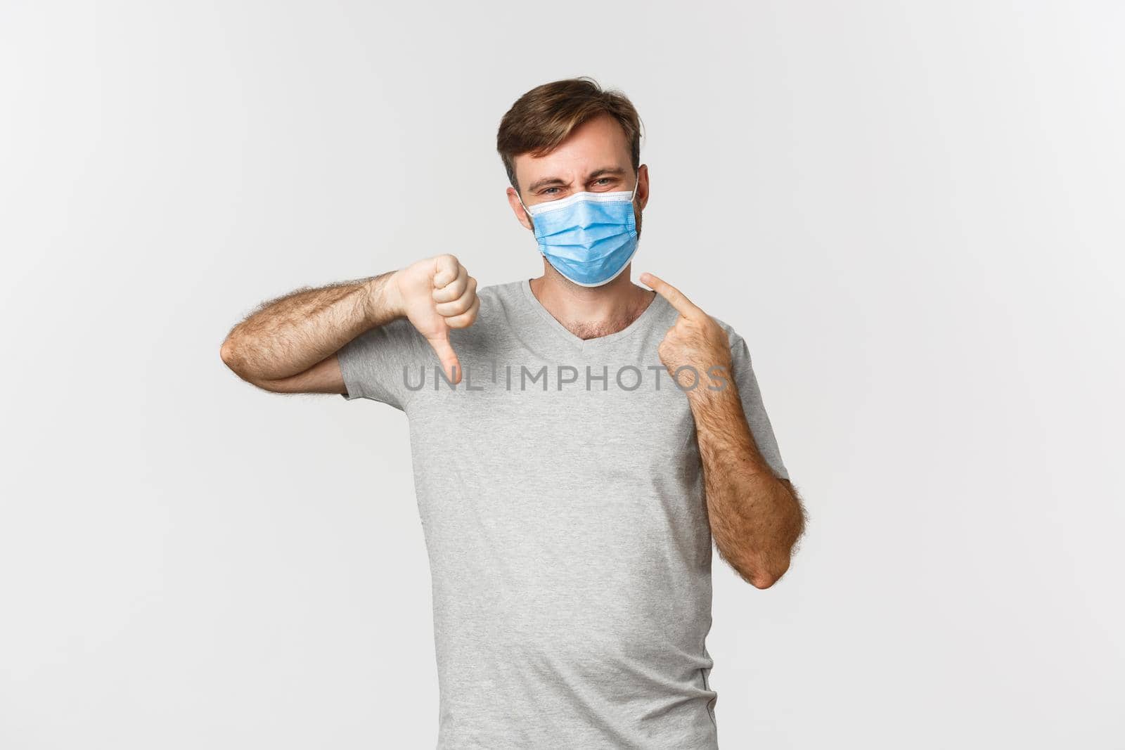 Concept of pandemic, covid-19 and social-distancing. Portrait of man hate wearing medical mask, complaining and showing thumbs-down, standing over white background.