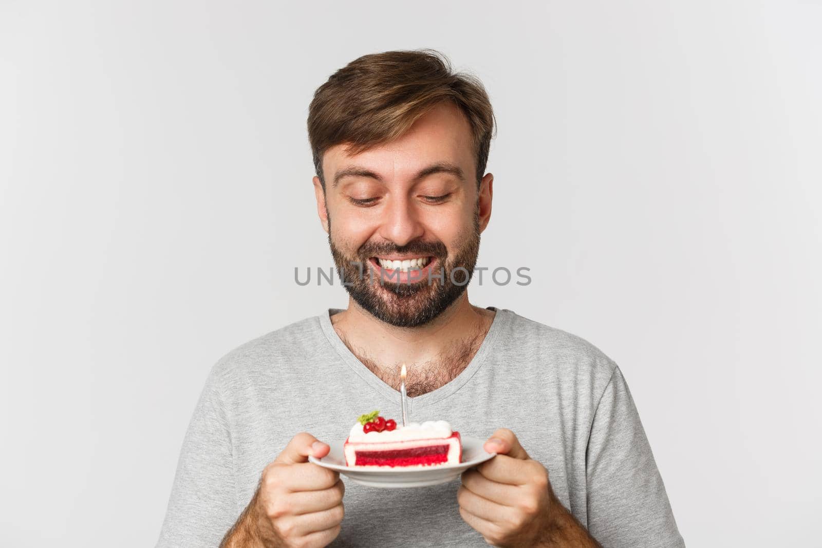 Close-up of happy bearded man, smiling and celebrating birthday, holding cake with lit candle, making b-day wish, standing over white background.