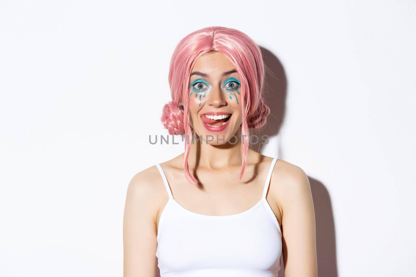 Close-up of attractive happy girl showing tongue and smiling, wearing pink party wig and bright makeup, standing over white background.