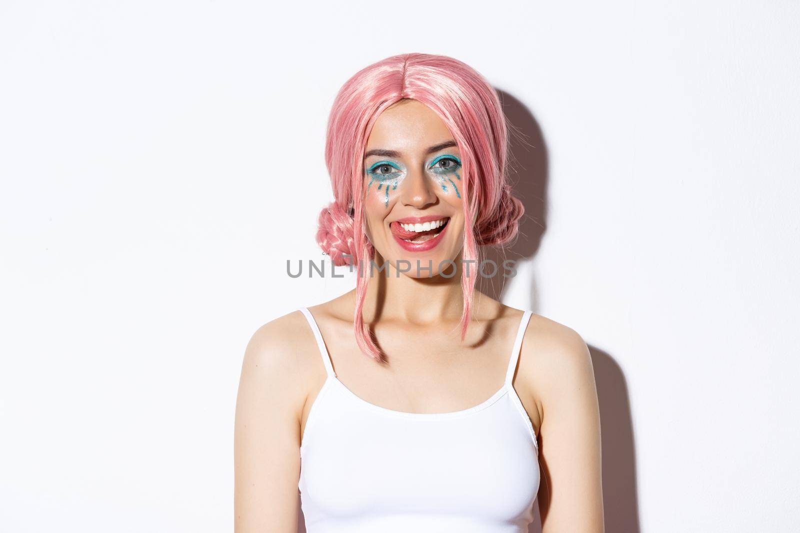 Close-up of happy beautiful female model in pink wig, showing tongue and smiling, celebrating holiday in fairy costume, standing over white background.