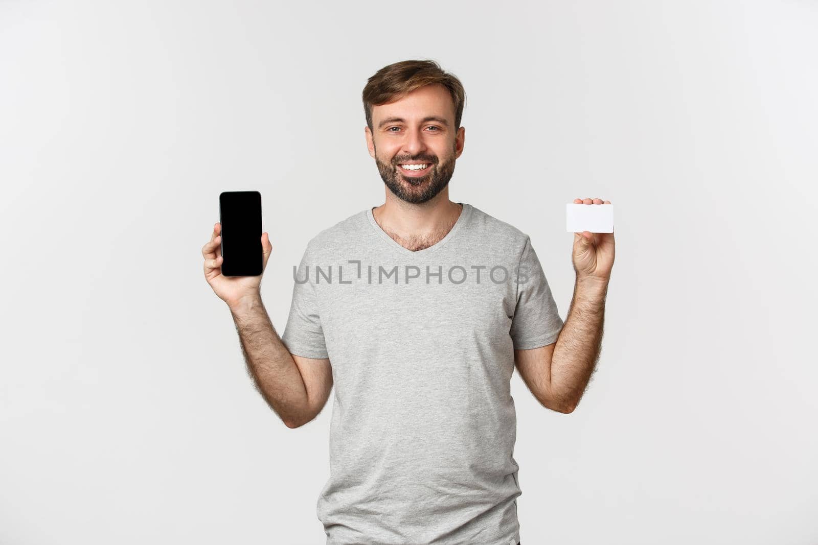 Handsome young man shopping online, holding credit card and mobile phone, showing smartphone screen, standing over white background.