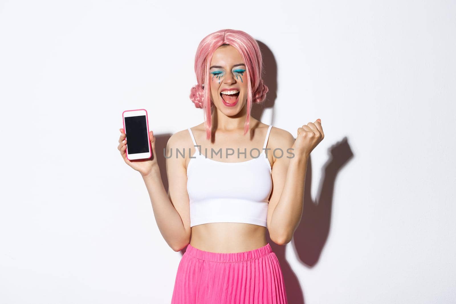 Image of beautiful party girl rejoicing, celebrating holiday, showing smartphone screen and shouting for joy, wearing pink wig and halloween outfit by Benzoix