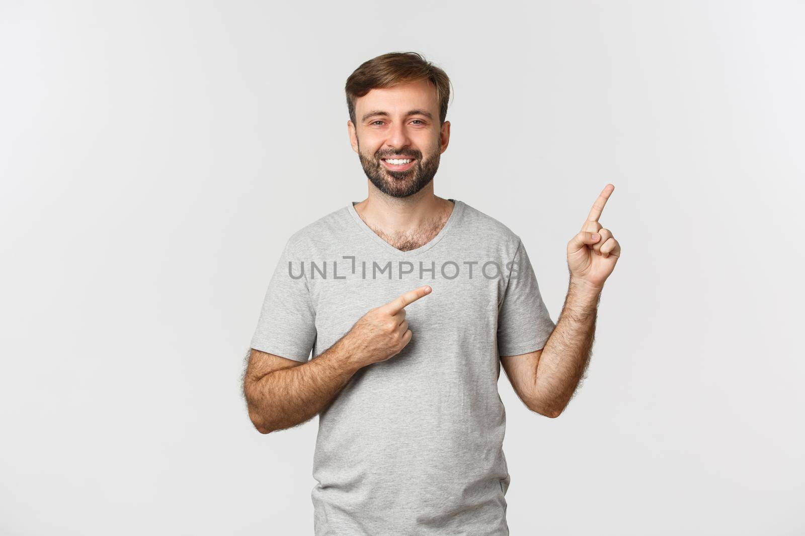 Image of happy handsome guy with beard, wearing gray t-shirt, pointing fingers at upper right corner logo and smiling, standing over white background by Benzoix
