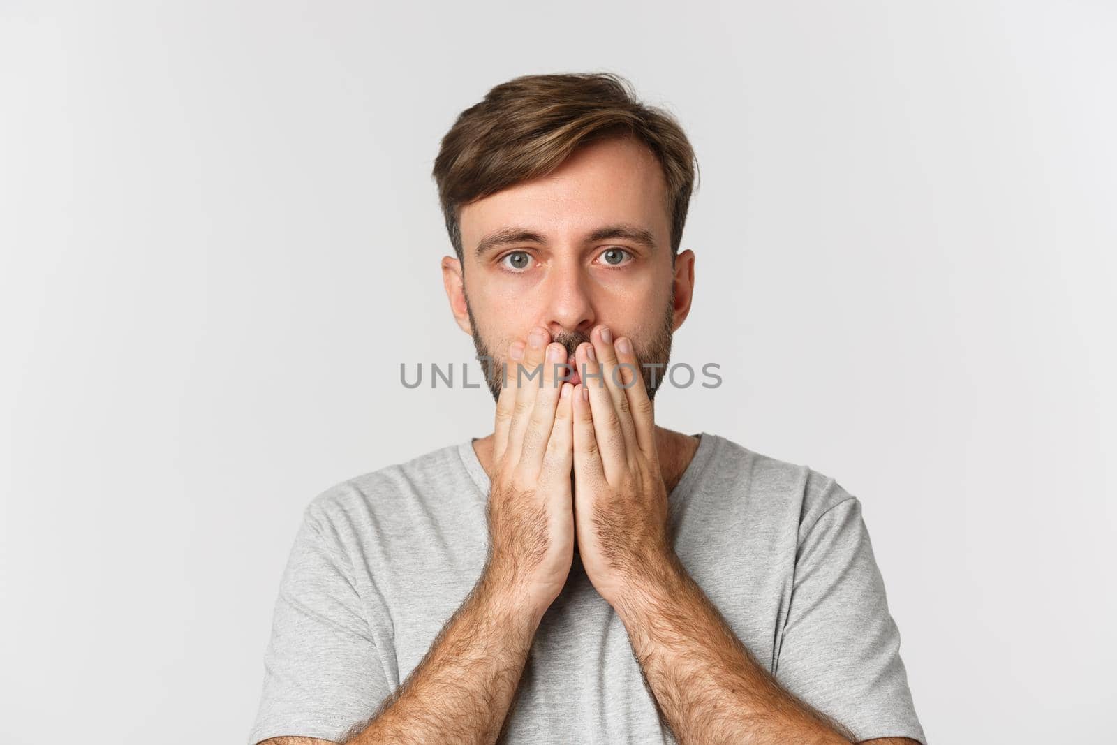 Close-up of shocked and speechless guy, cover mouth and looking at camera, standing over white background.