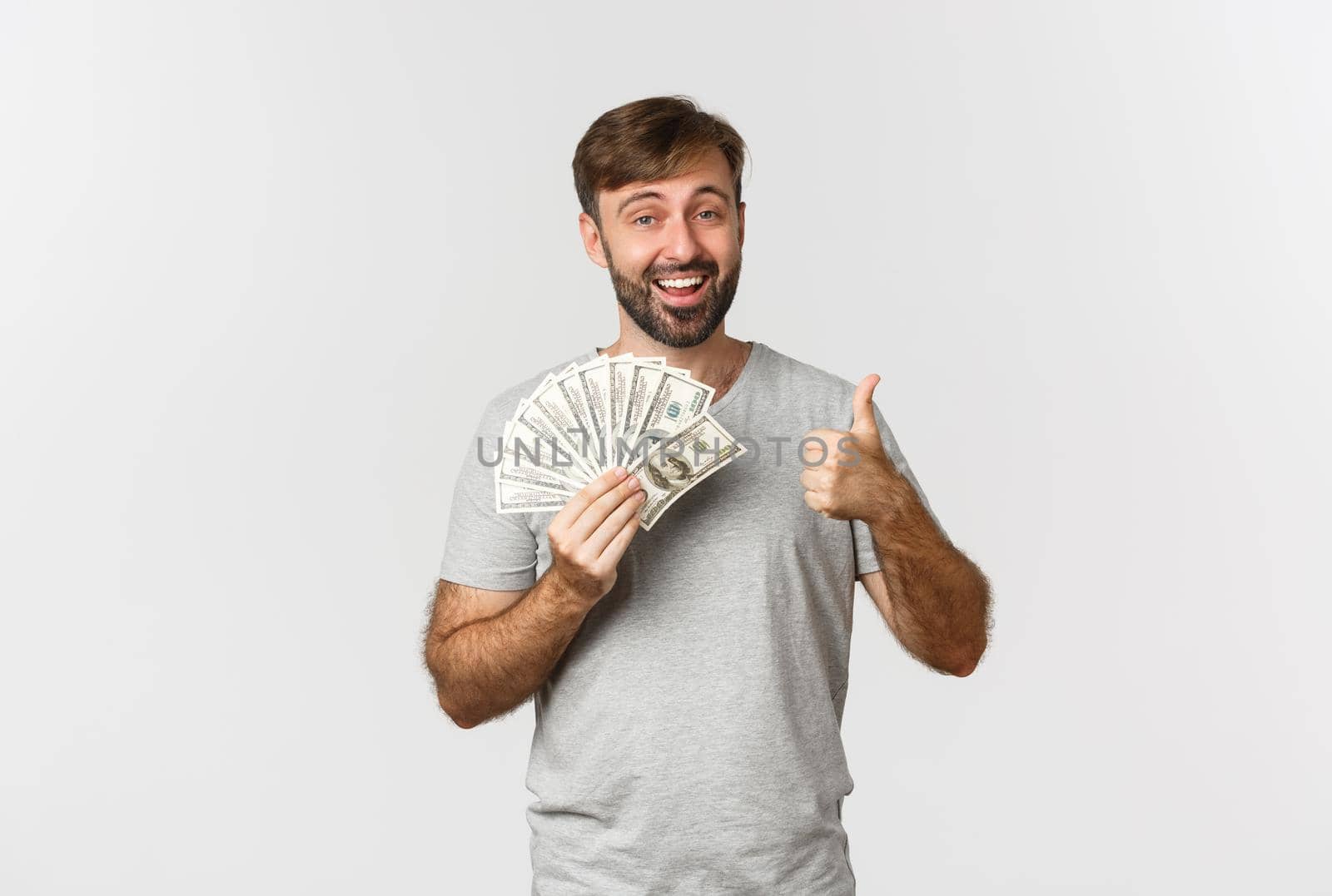 Portrait of handsome man with beard, showing thumbs-up and holding money, recommend credit or loan, standing over white background by Benzoix
