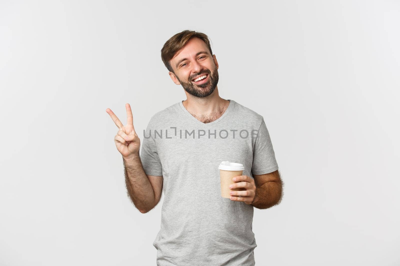 Portrait of carefree smiling man in gray t-shirt, drinking coffee and showing peace sign, standing over white background.