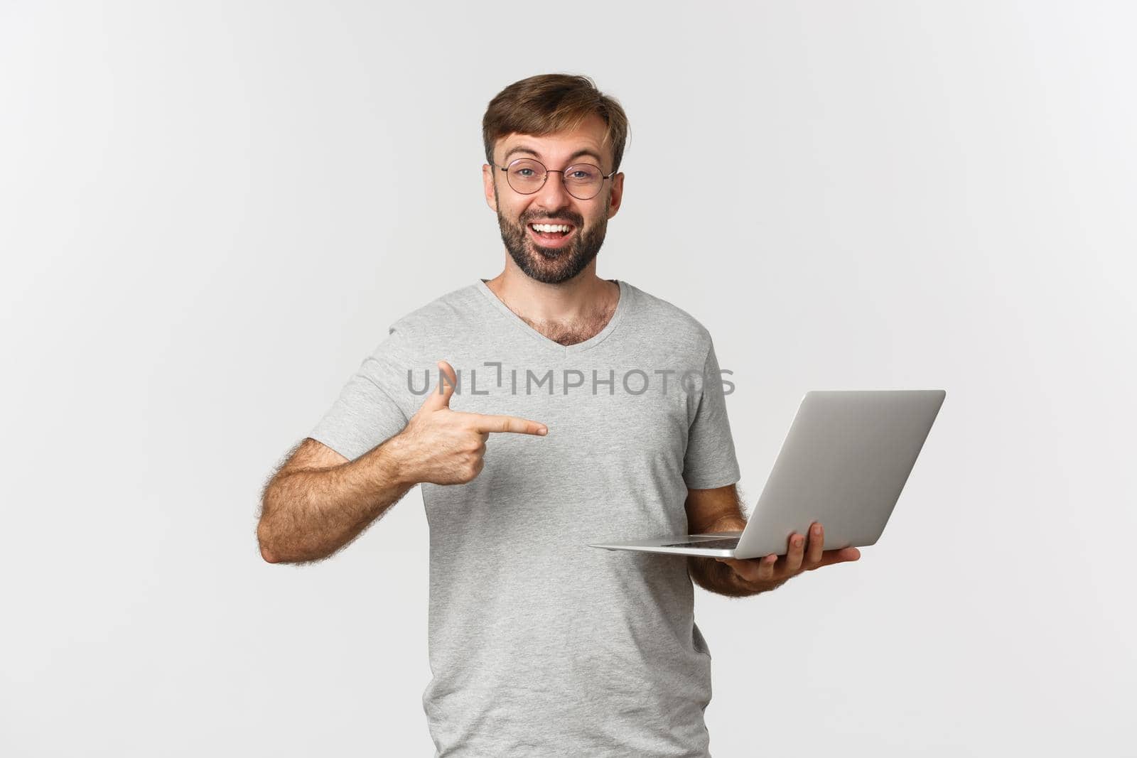Image of handsome man with beard, wearing glasses and gray t-shirt, pointing at laptop and smiling, recommend something, standing over white background.